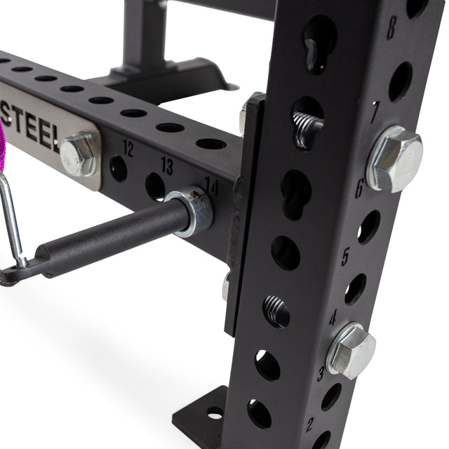 Close-up of a black Bells of Steel power rack with numbered holes (1–14), bolts, and Band Pegs with Carabiners attached by pin and clip. Partial "STEEL" label visible—perfect for resistance training or adding band pegs.