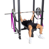 A user performs a bench press on a bench in a power rack, using Bells of Steel Band Pegs with Carabiners to attach purple resistance bands for added resistance during their workout.
