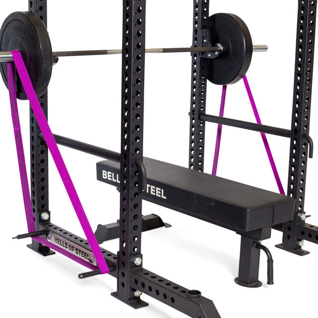 A black bench labeled "BELLS OF STEEL" is set in a power rack with a loaded barbell. Purple resistance bands are attached from Bells of Steel Standard Band Pegs at the rack’s base to each end of the barbell.