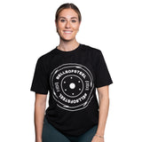A smiling woman wears a black Bamboo T-Shirt by Bells of Steel, featuring a large white weight plate graphic with "BELLS OF STEEL" and "25 KG" text on the front, standing against a plain white background.
