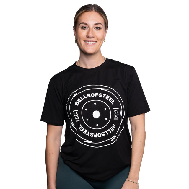 A smiling woman wears a black Bamboo T-Shirt by Bells of Steel, featuring a large white weight plate graphic with "BELLS OF STEEL" and "25 KG" text on the front, standing against a plain white background.