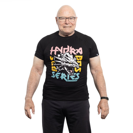 A smiling older man with glasses wears a black Bells of Steel Bamboo T-Shirt featuring colorful "Hydra Beast Series" text and a dragon graphic, standing against a white background.