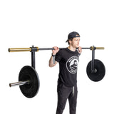 A person stands on a plain white background, wearing black clothes and a backwards cap, holding the Bells of Steel Camber Bar Attachment - Pair across their shoulders, preparing for a squat exercise.
