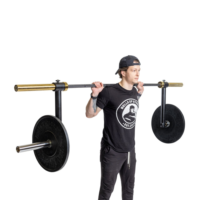 A person stands on a plain white background, wearing black clothes and a backwards cap, holding the Bells of Steel Camber Bar Attachment - Pair across their shoulders, preparing for a squat exercise.