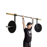 A person wearing a black cap and t-shirt lifts a Bells of Steel Camber Bar Attachment - Pair with two large, black weight plates featuring gold accents overhead against a white background.