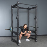 A woman in athletic wear performs a squat inside a power rack, using the Bells of Steel Safety Squat Bar – The SS4 with weights and a protective pad, in a gym with gray walls and flooring, highlighting the intensity of leg day.