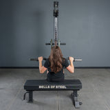 A person with long brown hair, seen from behind, uses the Bells of Steel Fat Bar - Olympic Curl Bar Cable Attachment for a lat pulldown, challenging their grip strength while seated in the gym.