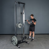 A man in a black t-shirt, shorts, and athletic shoes performs a bicep curl with the Bells of Steel Fat Bar - Olympic Curl Bar Cable Attachment on a cable machine in a gym with gray walls and a tiled floor.