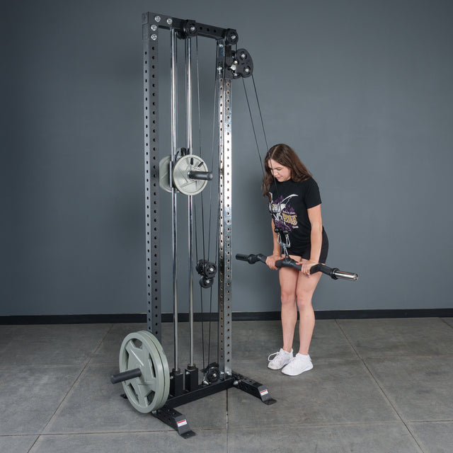 A woman in a gym with gray walls uses the Bells of Steel Fat Bar - Olympic Curl Bar Cable Attachment, gripping it with both hands to boost her grip strength as she stands ready to exercise.
