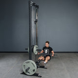 A person sits on a gym floor, holding the Bells of Steel Fat Bar - Olympic Curl Bar Cable Attachment on a tall cable machine with weight plates, preparing for a seated row. The background has plain gray walls and flooring.