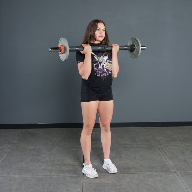 A woman in a black t-shirt and shorts stands indoors on a gray floor, holding a Bells of Steel Fat Bar - Olympic Curl Bar Cable Attachment at her shoulders as if preparing for a front squat or overhead press.