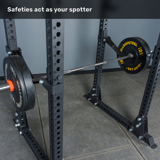 A Bells of Steel Manticore Flat Foot Power Rack - Prebuilt (3" x 3", 1" Holes) features sturdy uprights and safety arms that securely hold a barbell with weight plates, showcasing an efficient gym equipment setup for your home gym. Text at the top reads: "Safeties act as your spotter.