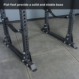 A Manticore Flat Foot Power Rack – Prebuilt (3" x 3", 1" Holes) by Bells of Steel, featuring flat feet and multiple adjustment holes, stands on a gray tiled floor. The text above reads, "Flat feet provide a solid and stable base." Perfect for any home gym, this rack offers durable 3x3 uprights for enhanced strength and reliability.