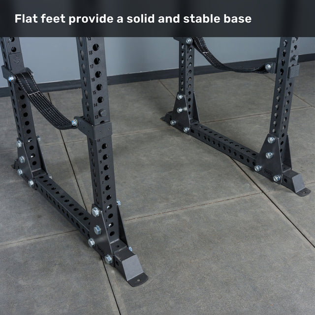 A Manticore Flat Foot Power Rack – Prebuilt (3" x 3", 1" Holes) by Bells of Steel, featuring flat feet and multiple adjustment holes, stands on a gray tiled floor. The text above reads, "Flat feet provide a solid and stable base." Perfect for any home gym, this rack offers durable 3x3 uprights for enhanced strength and reliability.