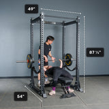 Two people use a black Bells of Steel Manticore Flat Foot Power Rack - Prebuilt (3" x 3", 1" Holes); one person is bench pressing while the other spots. The rack’s dimensions—49" wide, 54" deep, and 87¼" tall—are clearly marked. The plain gray wall and floor provide a minimalist backdrop, making this an ideal focused home gym setting.