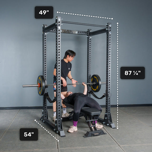 Two people use a black Bells of Steel Manticore Flat Foot Power Rack - Prebuilt (3" x 3", 1" Holes); one person is bench pressing while the other spots. The rack’s dimensions—49" wide, 54" deep, and 87¼" tall—are clearly marked. The plain gray wall and floor provide a minimalist backdrop, making this an ideal focused home gym setting.