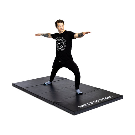 A person in black athletic wear does a wide-legged stance with arms extended on the Bells of Steel Four Fold Mat, set against a white background.