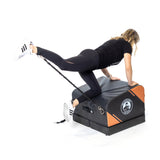 A woman in black athletic wear and white sneakers performs a resistance band leg kickback on the Bells of Steel Soft Glute Bench with black and orange accents.