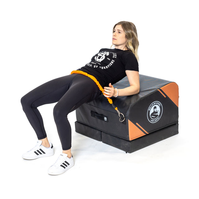 A woman in athletic wear does hip thrusts on the Bells of Steel Soft Glute Bench, using a resistance band around her hips. Her feet are flat on the floor and she looks straight ahead during the exercise.