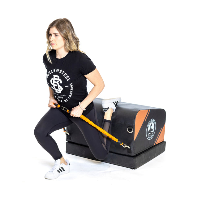 A woman demonstrates a split squat with a yellow resistance band, placing one foot on the Bells of Steel Soft Glute Bench. She sports a black t-shirt, black leggings, and white sneakers.