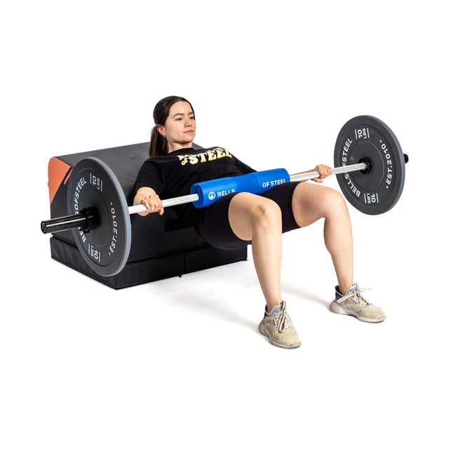 A woman performs a barbell hip thrust, resting her upper back on the Bells of Steel Soft Glute Bench and holding a barbell across her hips, with her feet flat on the floor.
