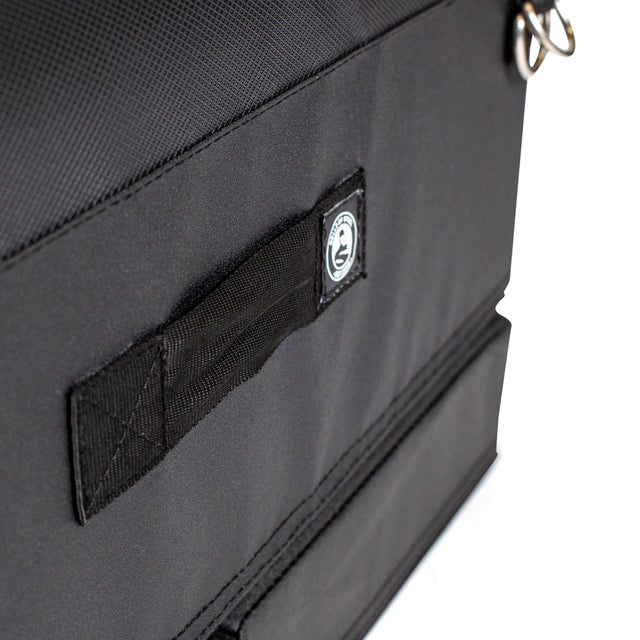 Close-up of a black fabric bag from Bells of Steel, featuring a stitched handle, small circular logo patch, and silver rings. The durable material is ideal for carrying your Soft Glute Bench or hip thrust bench accessories.