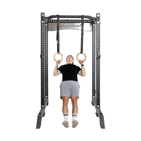 A person in a black shirt and gray shorts demonstrates strength and balance by performing a core workout on Bells of Steel Gymnastics Rings attached to a sturdy metal frame, keeping their feet off the ground to showcase the effectiveness of bodyweight training.