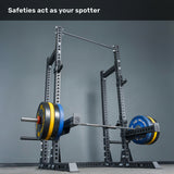 A loaded barbell rests on the safety arms of the Bells of Steel Manticore Half Rack - Prebuilt (3" x 3", 1" Holes) in a home gym, with the text "Safeties act as your spotter" displayed at the top.