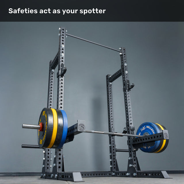 A loaded barbell rests on the safety arms of the Bells of Steel Manticore Half Rack - Prebuilt (3" x 3", 1" Holes) in a home gym, with the text "Safeties act as your spotter" displayed at the top.