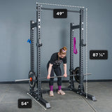 A woman prepares to lift a barbell inside the Bells of Steel Manticore Half Rack - Prebuilt (3" x 3", 1" Holes), a durable power rack ideal for any home gym, with labeled dimensions of 49" wide, 54" deep, and 87¼" tall. Resistance bands hang on the rack.