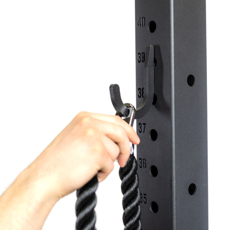 A hand hangs a black twisted rope cable attachment with a metal clip onto a Bells of Steel Utility Hook mounted on a vertical exercise rack marked with numbered holes—a smart gym storage solution.