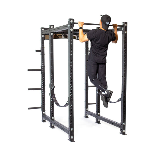 A person wearing athletic attire and a cap executes a pull-up on the Bells of Steel Hydra Six Post Power Rack - Prebuilt (3" x 3", ⅝" Holes), utilizing resistance bands and Hydra Attachments. The modular components of this power rack create a striking contrast against the pristine white background.