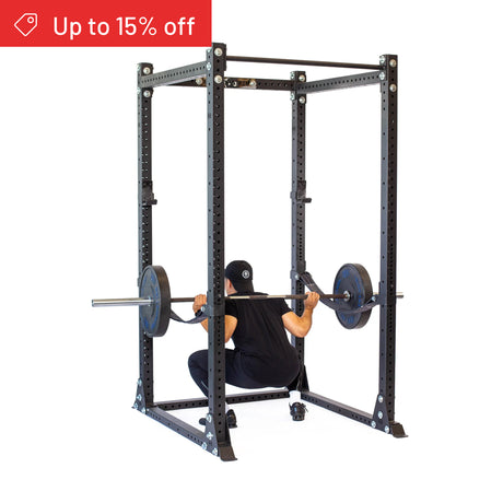 A person squats with a barbell loaded with plates inside the Bells of Steel Hydra Flat Foot Power Rack Builder (3" x 3", ⅝" holes). A red “Up to 15% off” banner appears in the corner. The plain white background suits any home gym.