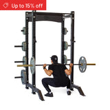 A person in black workout gear squats with a loaded barbell inside the Bells of Steel Hydra Collegiate Power Rack Builder (3" x 3", ⅝" Holes), with weight plates stored on the rack. A red banner says "Up to 15% off.