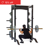 Wearing black athletic wear, a person does a barbell squat in the Bells of Steel Hydra Collegiate Power Rack Builder (3" x 3", ⅝" holes) loaded with weight plates. A red "10% off" banner is in the top left on a white background.