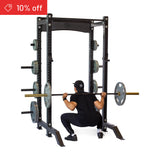 A person in black workout clothes performs a barbell squat in a Bells of Steel Hydra Collegiate Power Rack Builder (3" x 3", ⅝" Holes) with weight plates. A red "10% off" label appears in the top left on a white background.