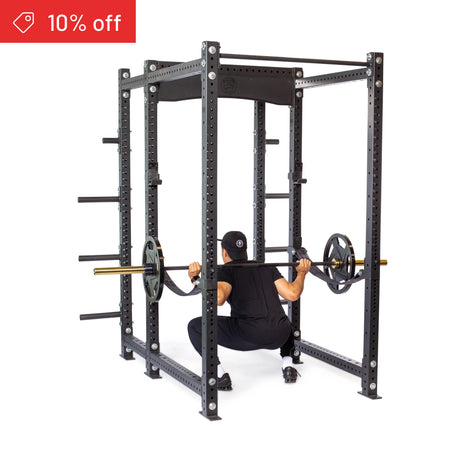 A person squats with a barbell inside the Bells of Steel Hydra Six Post Power Rack Builder (3" x 3", ⅝" Holes), wearing black. The black rack has weight plate storage pegs and a “10% off” label in the top left corner.
