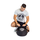 A man in a grey Bells of Steel t-shirt kneels while adjusting a large black Bells of Steel Fitness Sandbag labeled "100 LBS" on a white background, highlighting its durable construction.
