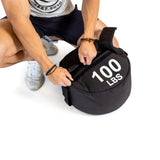 Wearing athletic gear, a person crouches to fasten the strap on a round black "Bells of Steel Fitness Sandbag" labeled "100 LBS," highlighting its durable construction against a plain white background.