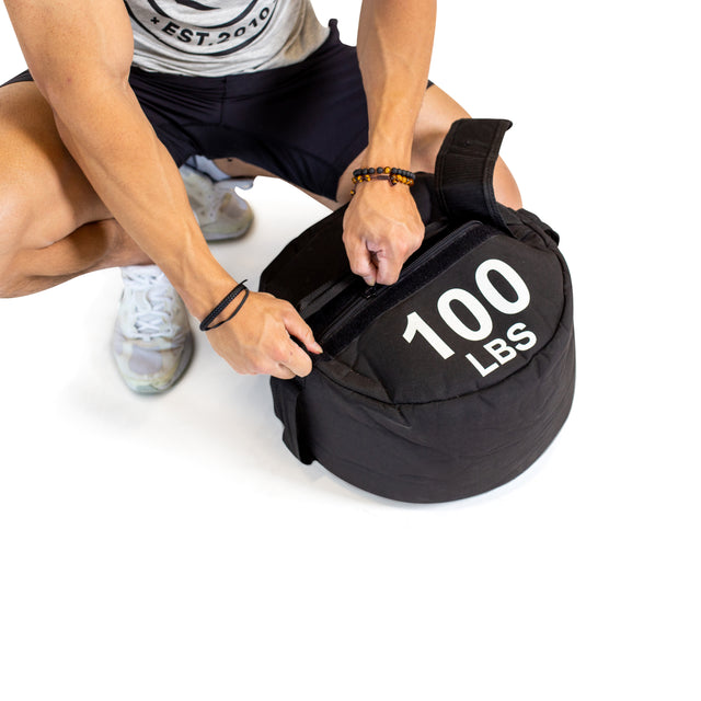 Wearing athletic gear, a person crouches to fasten the strap on a round black "Bells of Steel Fitness Sandbag" labeled "100 LBS," highlighting its durable construction against a plain white background.