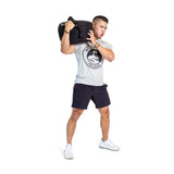 A man in a gray t-shirt, black shorts, and white sneakers lifts a Bells of Steel Fitness Sandbag onto his shoulder with bent knees against a white background, showcasing the versatility of Fitness Sandbags.