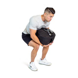 A man in a light gray T-shirt, black shorts, and white sneakers squats while lifting Bells of Steel Fitness Sandbags with durable Condura construction, staying focused on his workout against a plain white background.