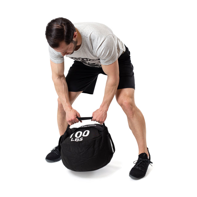 A man in a gray T-shirt and black shorts bends to grip the handles of a Bells of Steel Fitness Sandbag labeled "100 LBS" against a white background, showcasing its versatility for strength training.