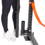A person in black leggings and white sneakers is holding a black barbell near gym equipment, which includes a power rack. An orange strap enhances the setup, featuring the Vertical Mount Barbell Holder Rack Attachment from Bells of Steel for efficient home gym organization. The individual confidently stands on a white surface.