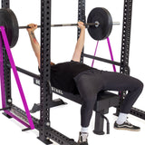 A person bench presses a barbell inside a power rack, using Bells of Steel Standard Band Pegs with resistance bands attached for support. Pink safety straps are in place. The lifter wears a black shirt, joggers, and sneakers.