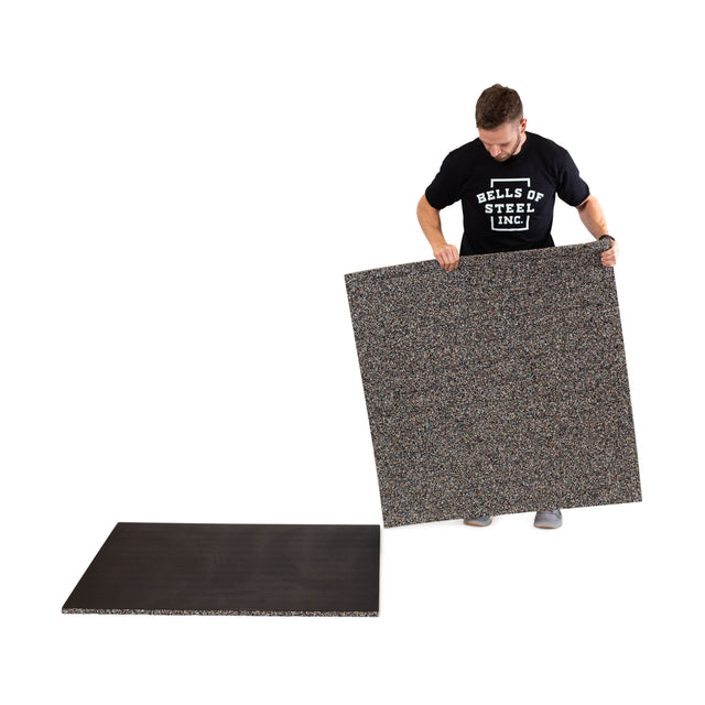 A man in a Bells of Steel t-shirt lifts a Rubber Flooring Gym Mat 39" x 39", known for sound dampening, while another Bells of Steel mat lies flat on the floor nearby.