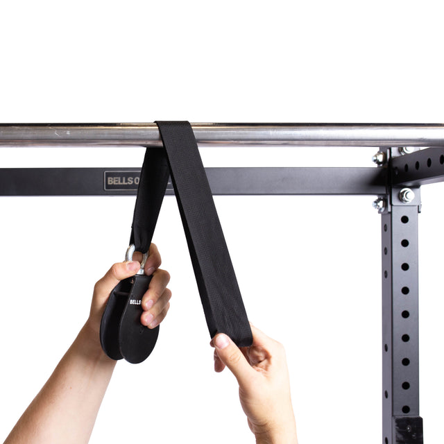 Two hands attach the black Bells of Steel Cable Pulley strap to a metal pull-up bar on a gym rack. This fitness accessory is ideal for tricep pushdowns or lat pulldowns, shown against a plain white background.