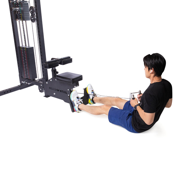A person in a black t-shirt and blue shorts uses the Bells of Steel Double D Handle Cable Attachment while performing a seated cable row on a weight machine, sitting with legs extended.