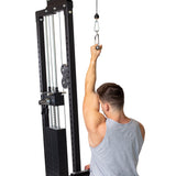 A person in a gray tank top uses a cable pulley machine, pulling a Bells of Steel Single D Handle Cable Attachment overhead with one arm extended. The machine features visible weight stacks, metal cables, and rubberized grips for comfort.