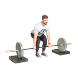 A man in a gray T-shirt and black pants prepares to deadlift a barbell resting on Bells of Steel Stackable Pull Blocks. He grips the bar with both hands, ready to lift, against a plain white background.